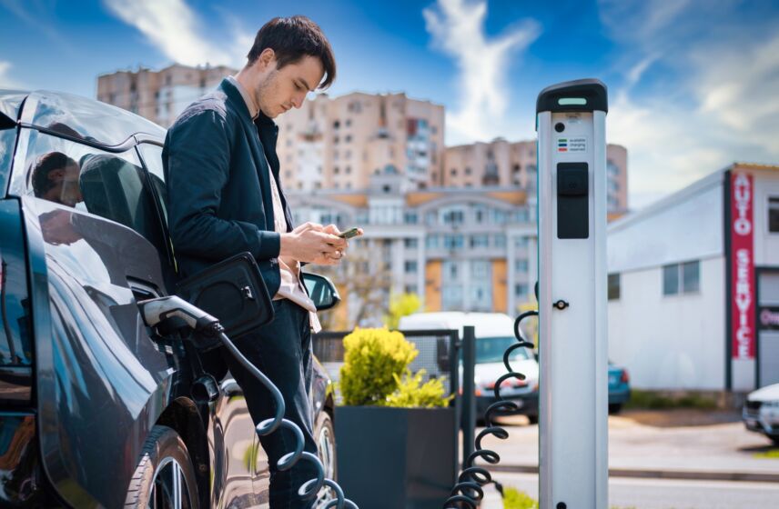 Man charging his electric car at charge station and using smartphone